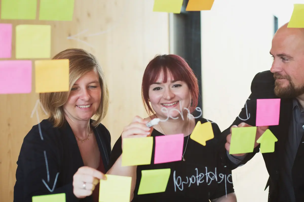 Zwei Frauen (links) und ein Mann (rechts) kleben Post-its mit Notizen an ein eine Scheibe.