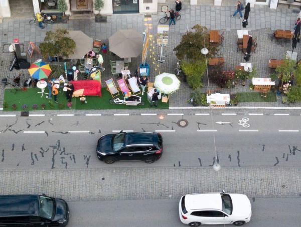 Vogelperspektive auf eine Straße und umgenutzte Parkplatzflächen. Auf der Straße fahren drei Autos.