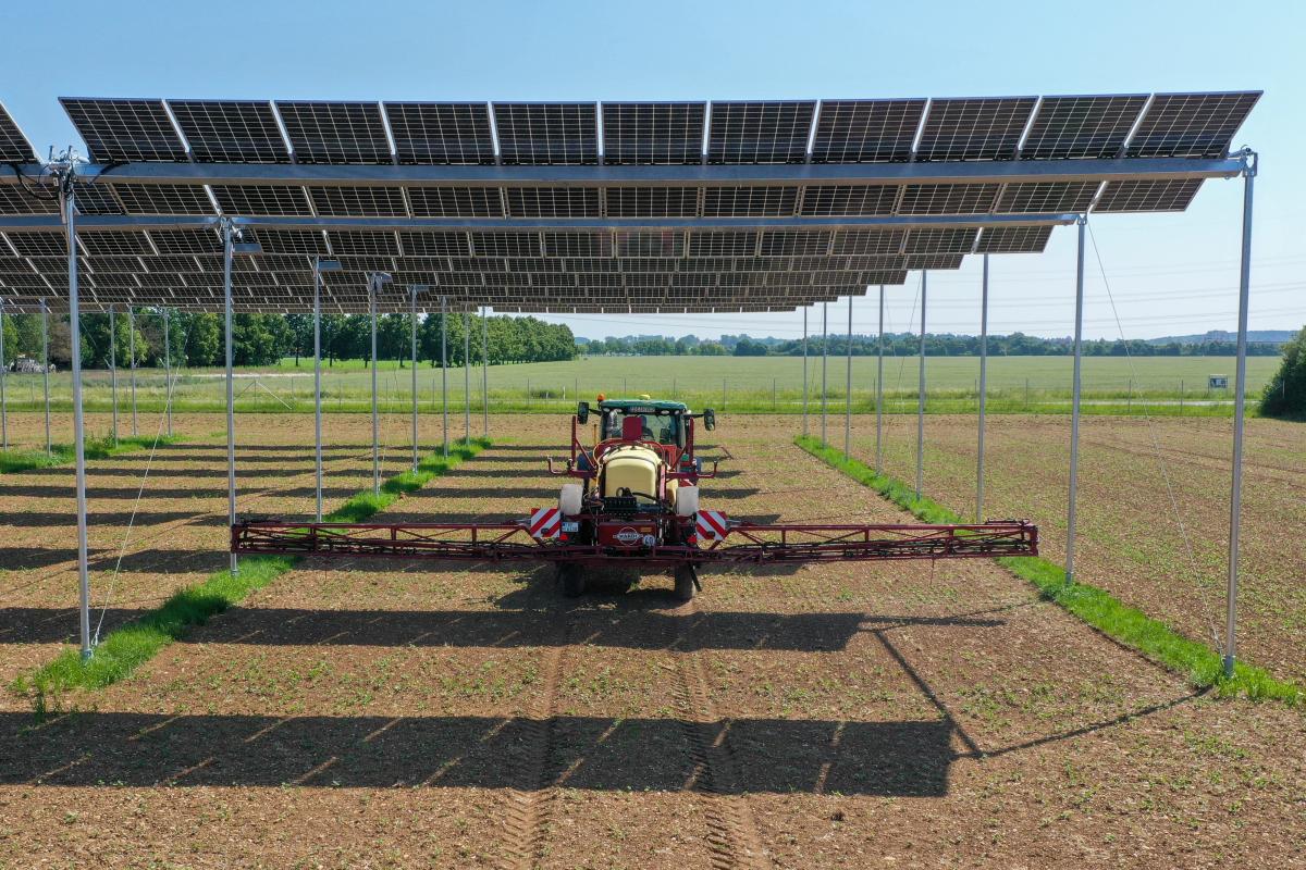 Traktor mit Feldspritze fährt über einen Acker unter erhöht montierten und beweglichen Solarmodulen, die der Sonne folgen. Der Himmel ist blau und wolkenlos.