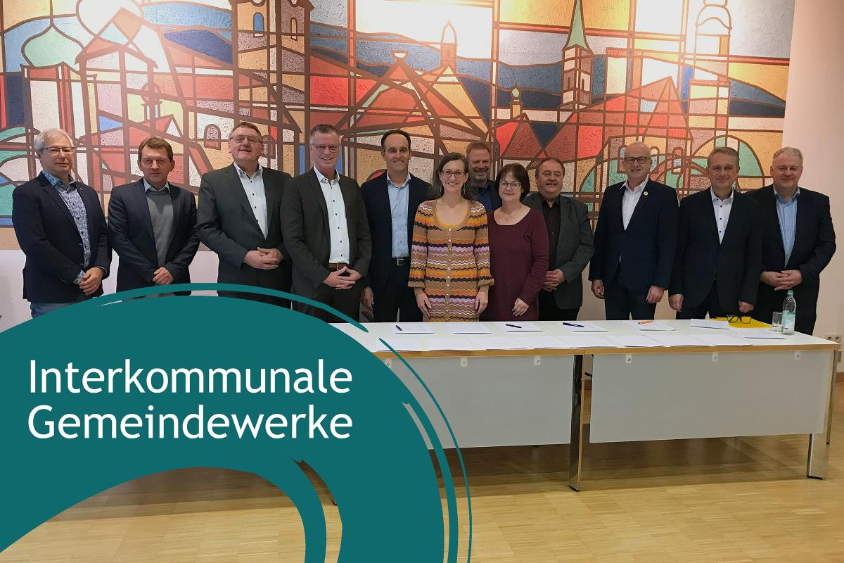 Personen stehen in einer Reihe vor einer bunten Wand und sehen lächelnd in die Kamera. Linke untere Ecke: Blauer Farbbanner mit dem Schriftzug "interkommunale Gemeindewerke"