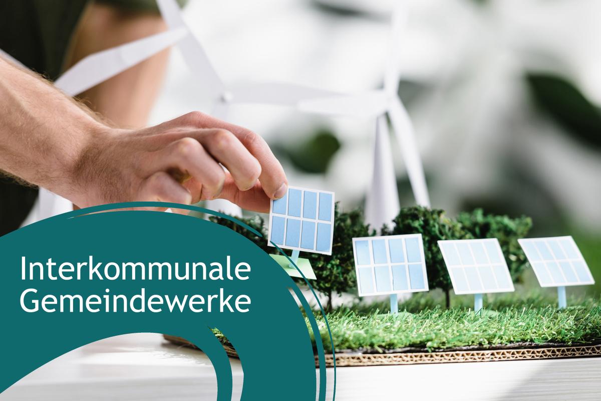 Im Hintergrund Hand, die Papier-PV-Anlage auf einem Modell platziert. Linke untere Ecke: Blauer Farbbanner mit dem Schriftzug "interkommunale Gemeindewerke"