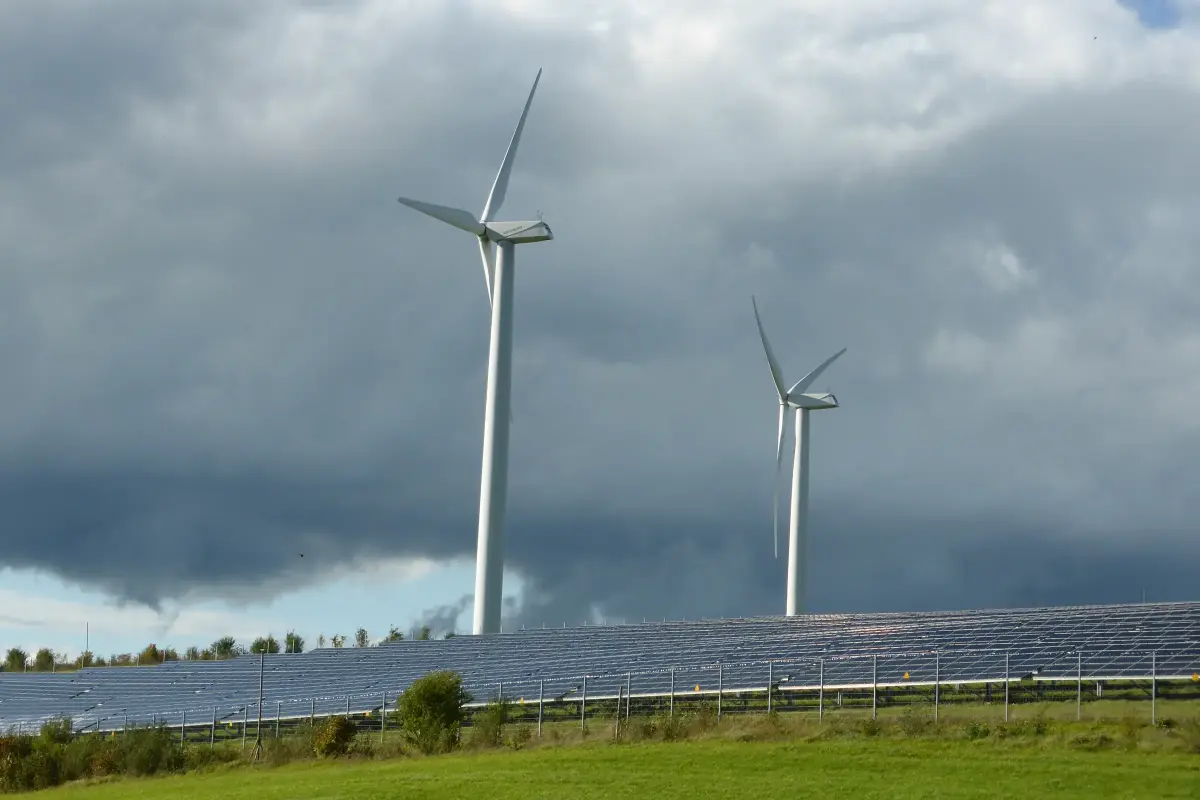Photovoltaik-Freiflächenanlage im Vordergrund und zwei Windkraftanlagen im Hintergrund bei bewölktem Himmel.