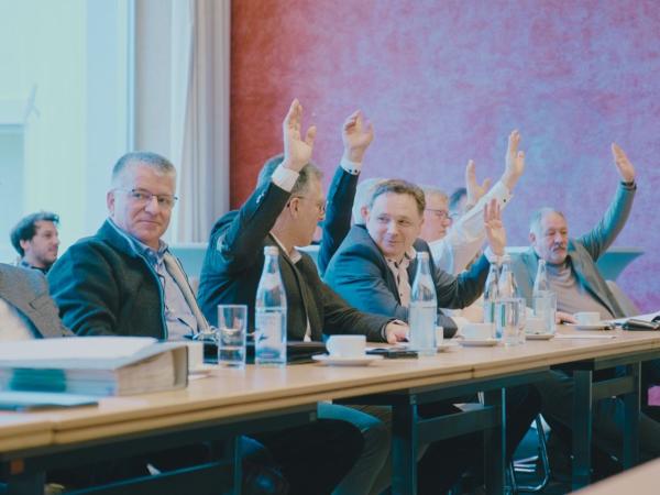 Männer sitzen an einem Tisch und heben Hände zur Abstimmung in die Luft.