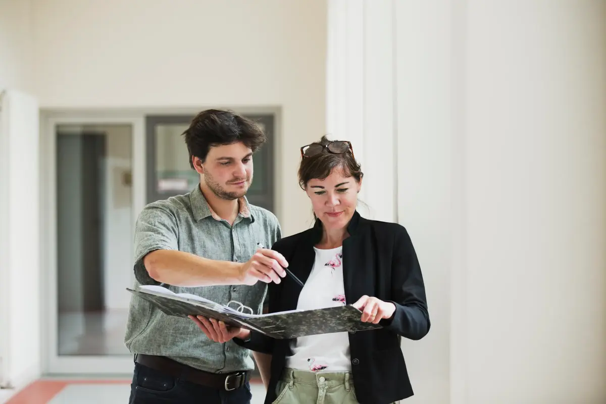 Eine Frau und ein Mann stehen in einem langgestreckten, hellen Flur mit hohen Decken zusammen. Die Frau hält einen aufgeschlagenen Aktenordner, in den sie gemeinsam blicken. Der Mann zeigt mit dem Stift auf eine aufgeschlagene Seite.