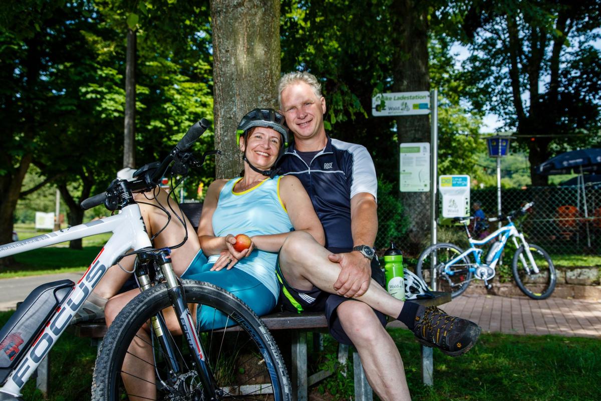 Mann und Frau sitzen auf Bank und lachen in die Kamera.