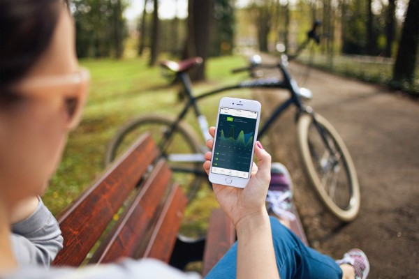 Frau sitzt auf Parkbank und hält Smartphone mit STADTRADELN-App in rechter hand. Im Hintergrund ein Fahrrad auf einem Fahrradweg.