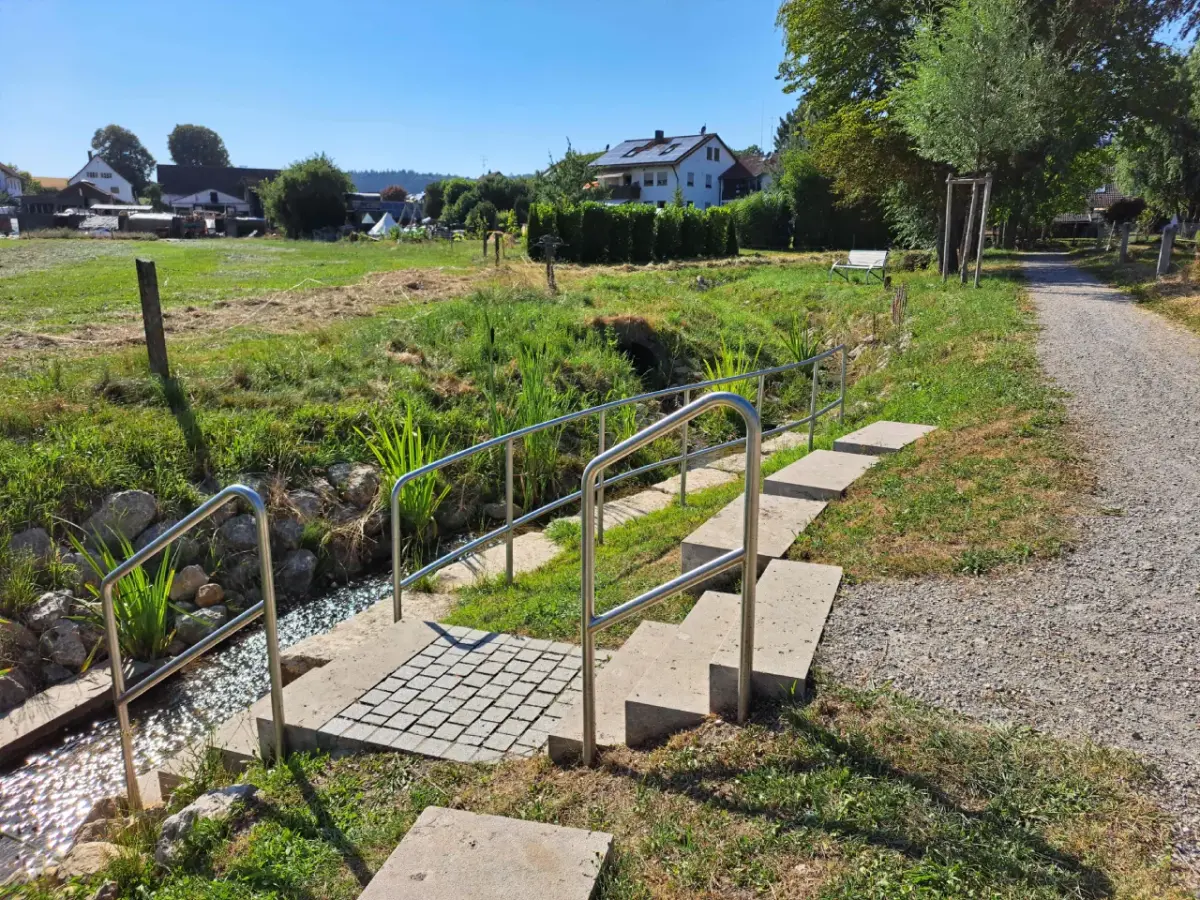 Renaturierter Bach mit Natursteinen, Bepflanzung und Treppe zum Bachlauf. Im Hintergrund Häuser bei gutem Wetter.