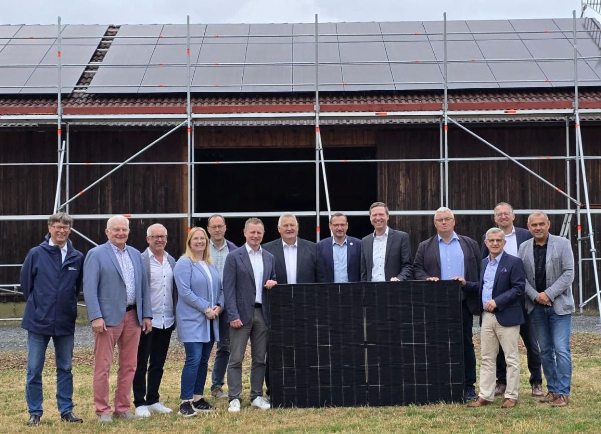 Gruppe hält PV Panel und steht vor einer Scheune mit PV auf dem Dach.