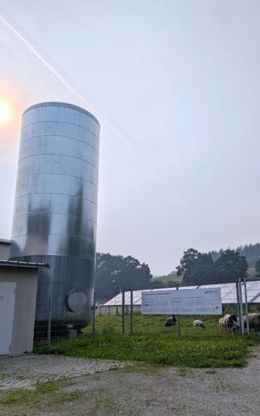 Der 100m³ große Pufferspeicher dient der Versorgungssicherheit | © Jana Schlesinger Links Pufferspeicher bei Sonnenschein, rechts PV-Freiflächenanlage.