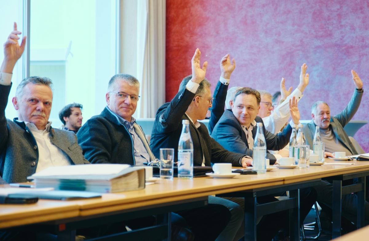 Männer sitzen an einem Tisch und heben Hände zur Abstimmung in die Luft.