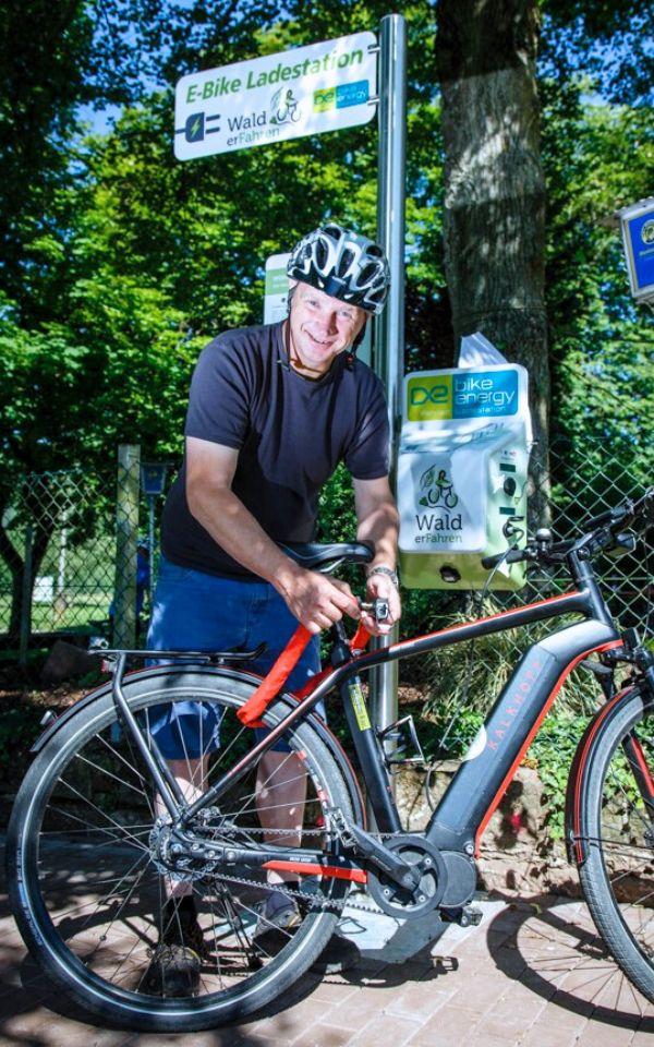 Man lehnt auf rotem Fahrrad. Im Hintergrund Schid zur E-Bike Ladestation.