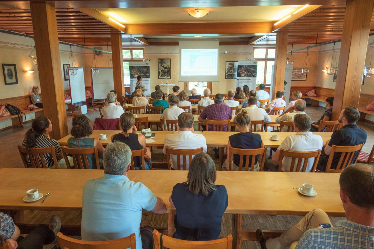 Menschen sitzen in Saal an Tischen und hören Vortrag zu.