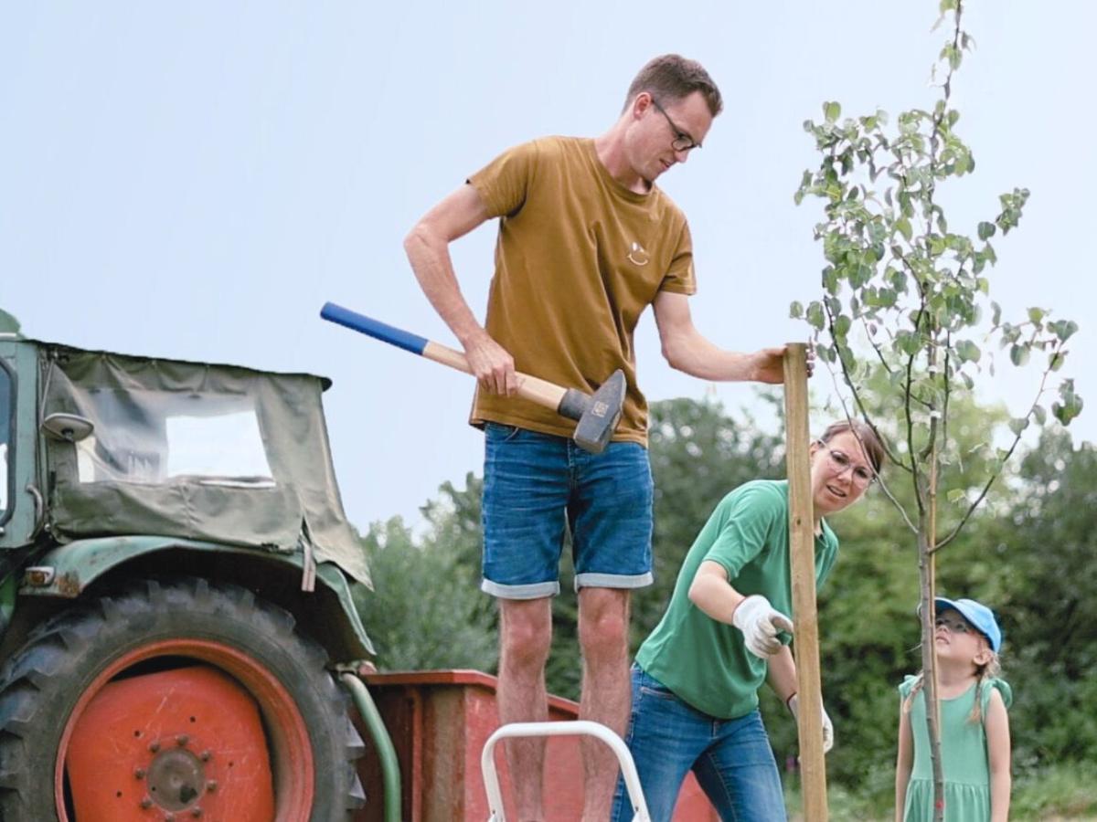 Links Traktor. Mitte Mann, rechts daneben Frau mit Tochter. Festbinden eines Baumes.