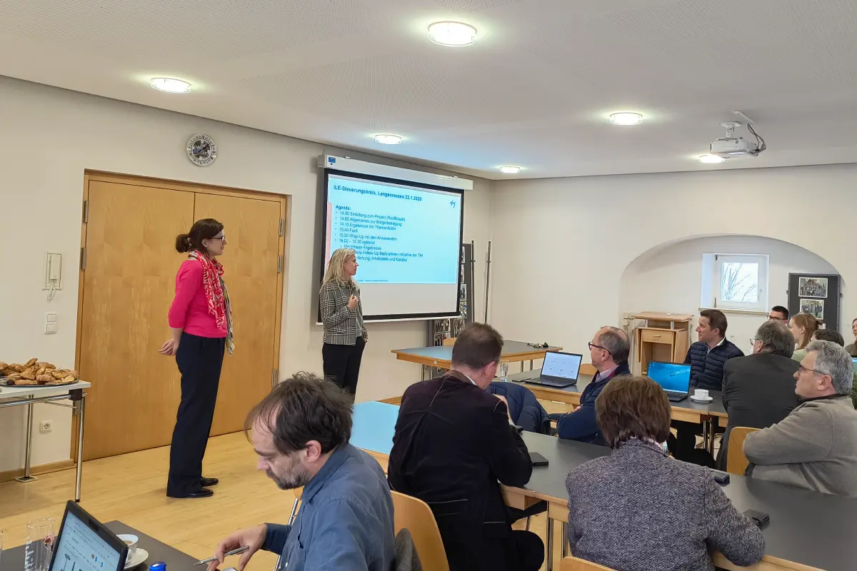 Zwei Professorinnen stehen vor einer Leinwand. Mehrere Personen sitzen an Tischen und sehen zur Leinwand.