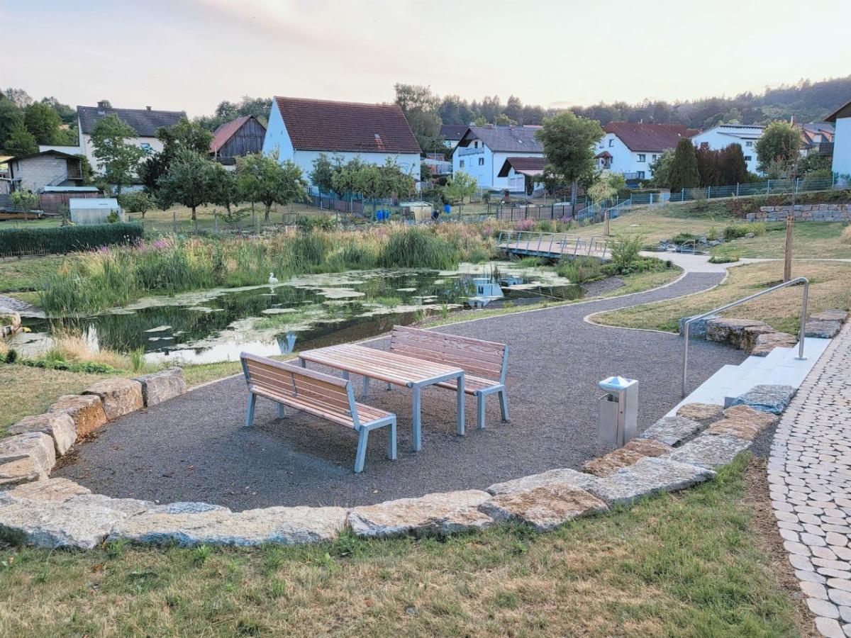 Vor dem Ortssaum von Waldfenster schmiegt sich ein kleiner See in den sanften Abhanf. Davor: Holzsitzgruppe auf einer dunkel geschotterten Fläche. Rechts: Wassertretbecken.