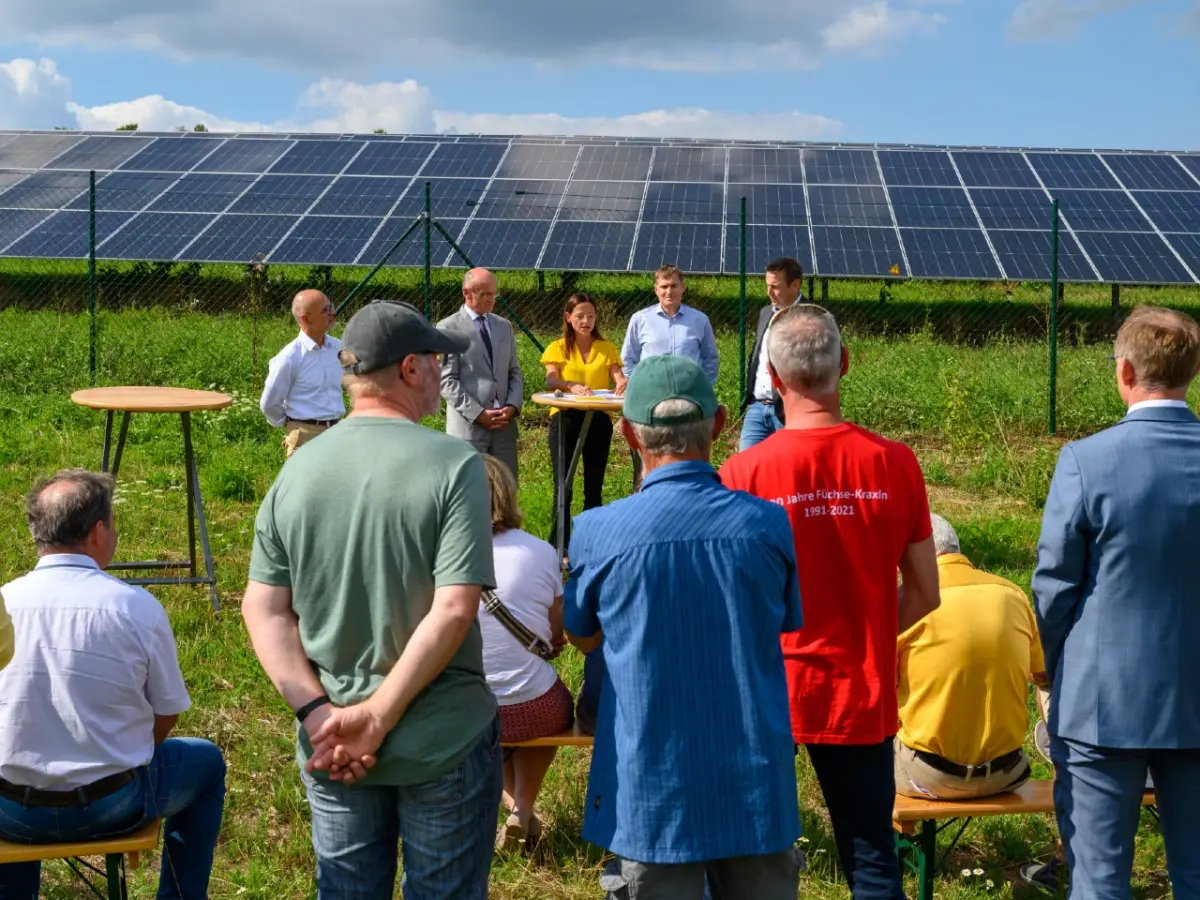 5 Personen stehen um Tisch und schauen richtugn Publikum, welches mit den Rücken zum Fotografen steht. Im Hintergrund: PV-Freiflächenanlage.