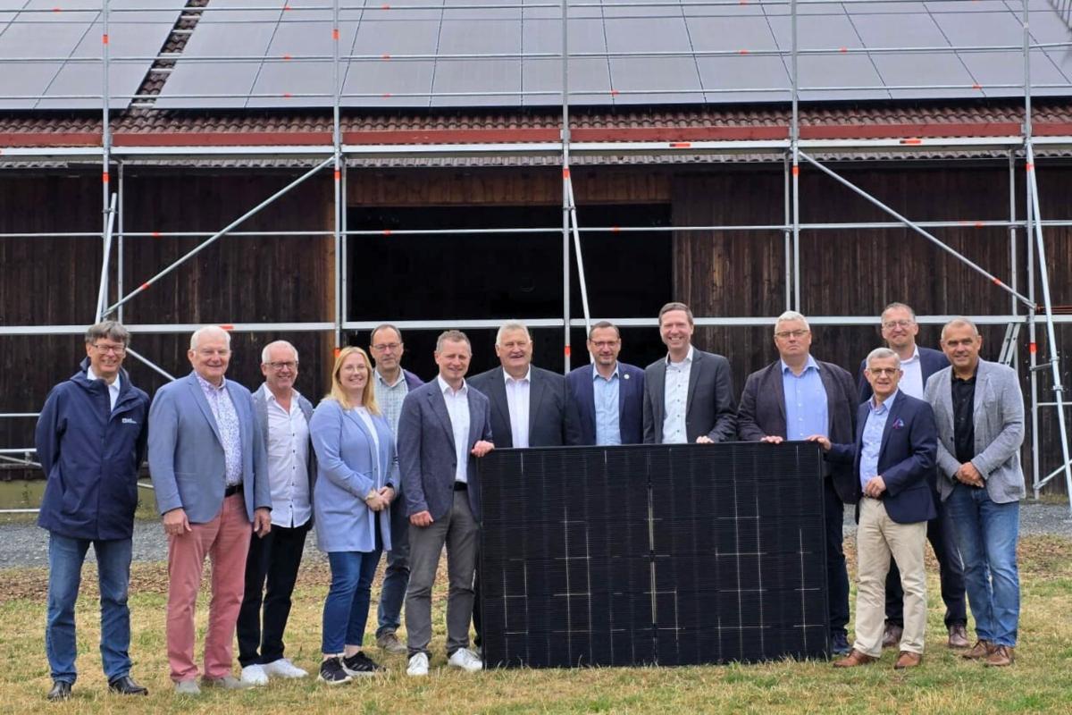 Gruppe hält PV Panel und steht vor einer Scheune mit PV auf dem Dach.