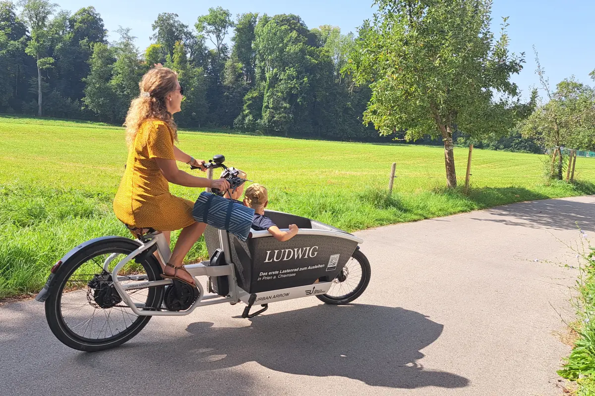 Frau in gelben Kleid fährt mit Lastenrad, in welchem zwei Kinder sitzen.