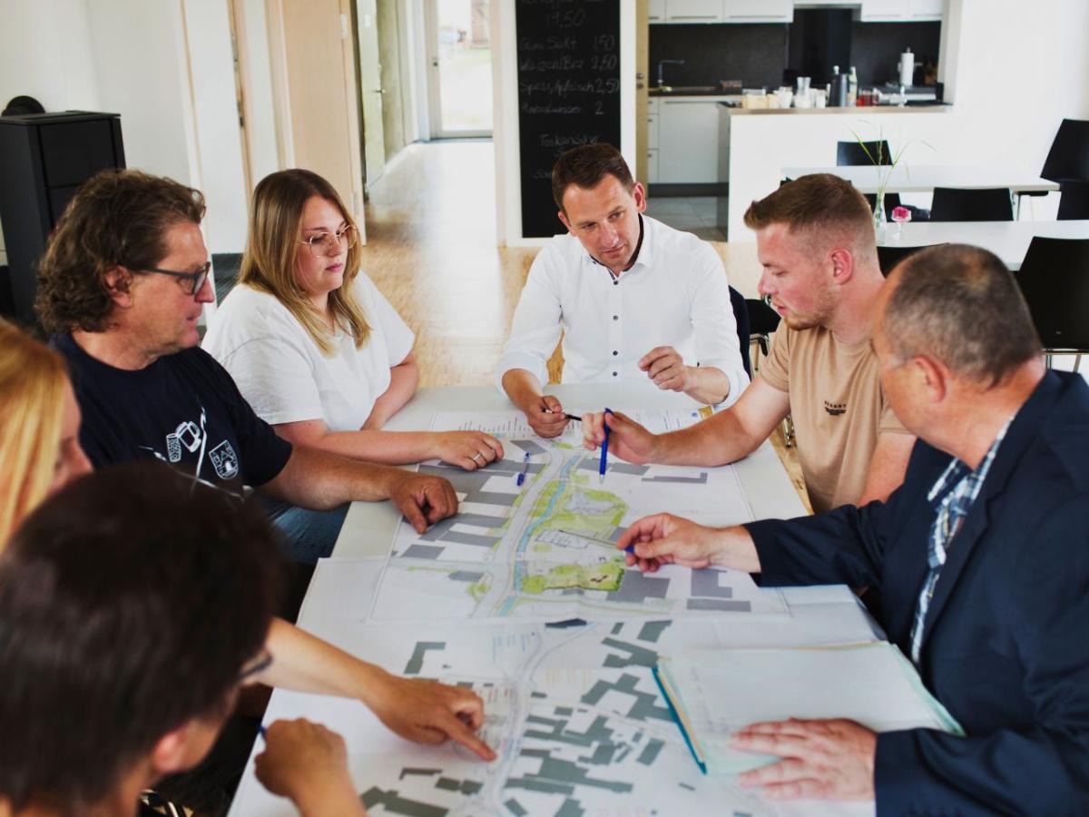 7 Personen sitzen an einem Besprechungstisch mit ausgebreiteten Entwurfsplänen zusammen und sprechen miteinander