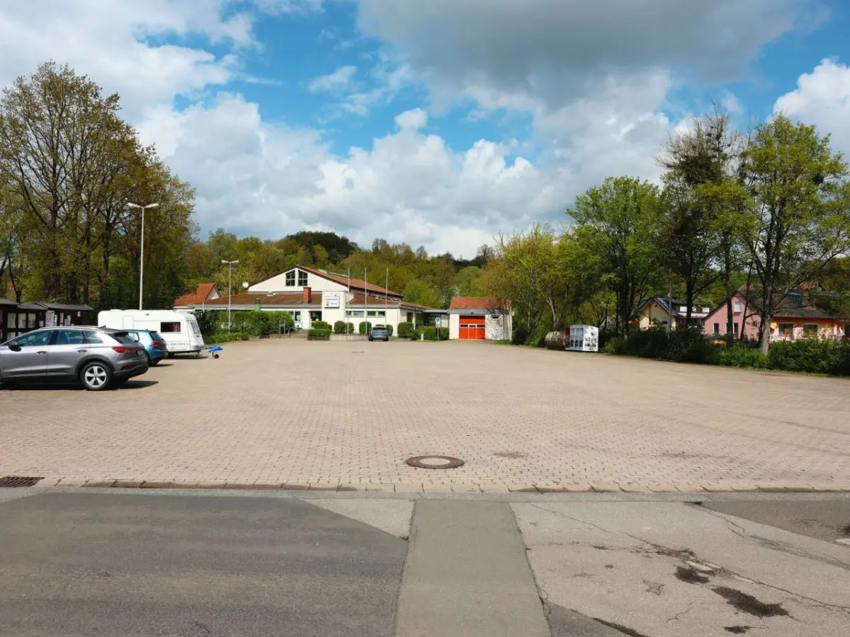 Gepflasterter Parkplatz. Zwei Autos und ein Wohnwagen abgestellt. Bäume säumen den Parkplatz. Im Hintergrund Häuser.