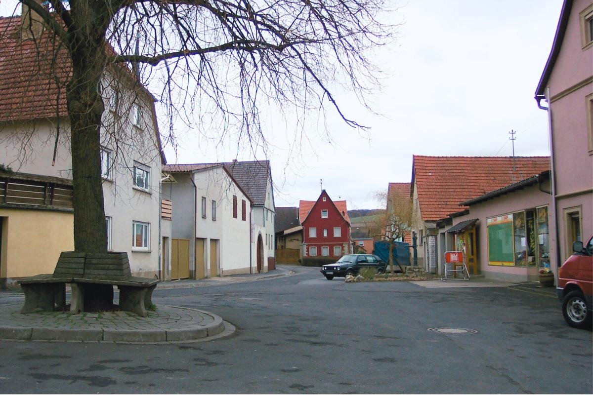 Asphaltierter Dorfplatz mit großer Linde und Sitzgelegenheiten im Zentrum.