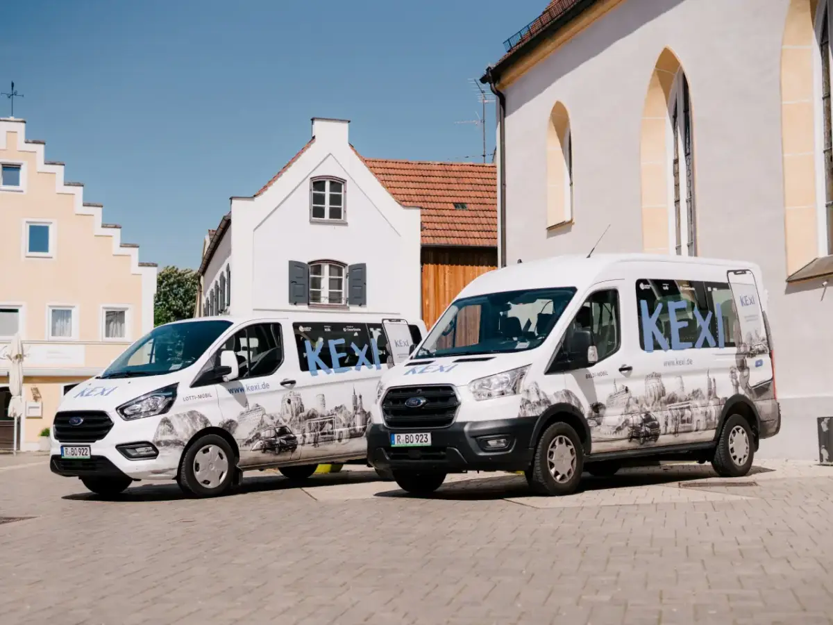 Zwei KEXI-Busse stehen auf Stadtplatz. Im Hintergrund Kirche und Häuser. Gutes Wetter.