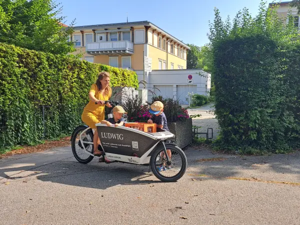 Frau in gelben Kleid fährt in einer Hofeinfahrt mit einem Lastenrad, in welchem zwei Kinder sitzen.