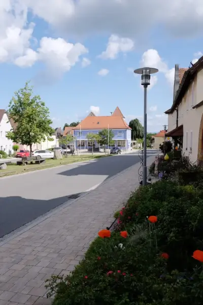 Rechts Blumenbeet. In der Mitte ein gepflasterter Gehweg. Im Hintergrund der Dorfplatz.