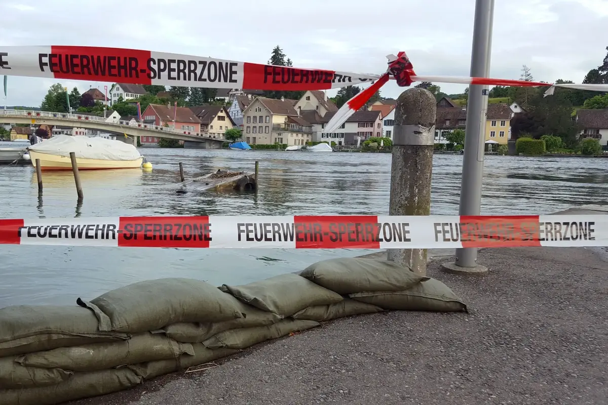 Fluss mit Hochwasser in Ortschaft. Snadsäcke und Feuerwehrabsperrband im Vordergrund.