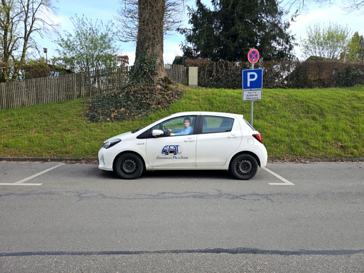 Frau sitzt in weißem Carsharing Auto, das auf einem Parkplatz für Carsharingsautos steht. Im Hintergrund ein Parkschild.