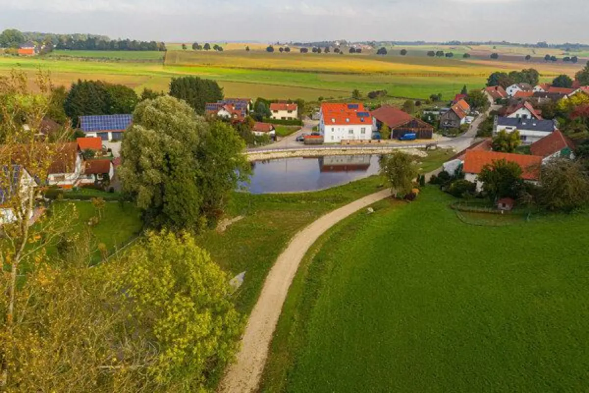 Weiher in Dorfmitte. Ein Weg führt im Vordergrund in das Dorf. Bäume links im Bild zu sehen.