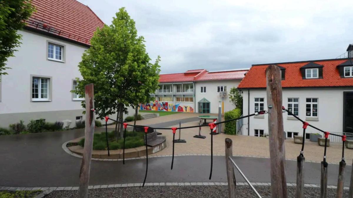 Schulhof im Hintergrund. Vorne Klettergerüst und Laubbaum.