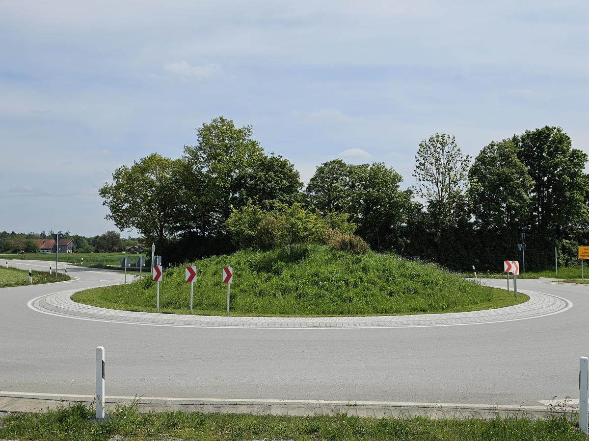 Begrünter Kreisverkehr um ländlichen Raum. Im Hintergrund Bäume und blauer Himmel.