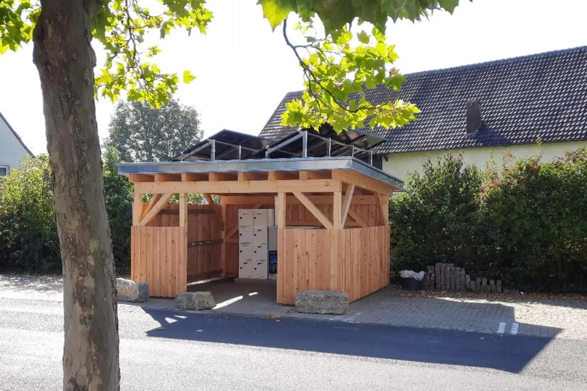 E-Bikeport aus Holzbauweise steht auf einem Parkplatz. Im Hintergrund eine Hecke und ein Haus. Im Vordergrund ein Baum.