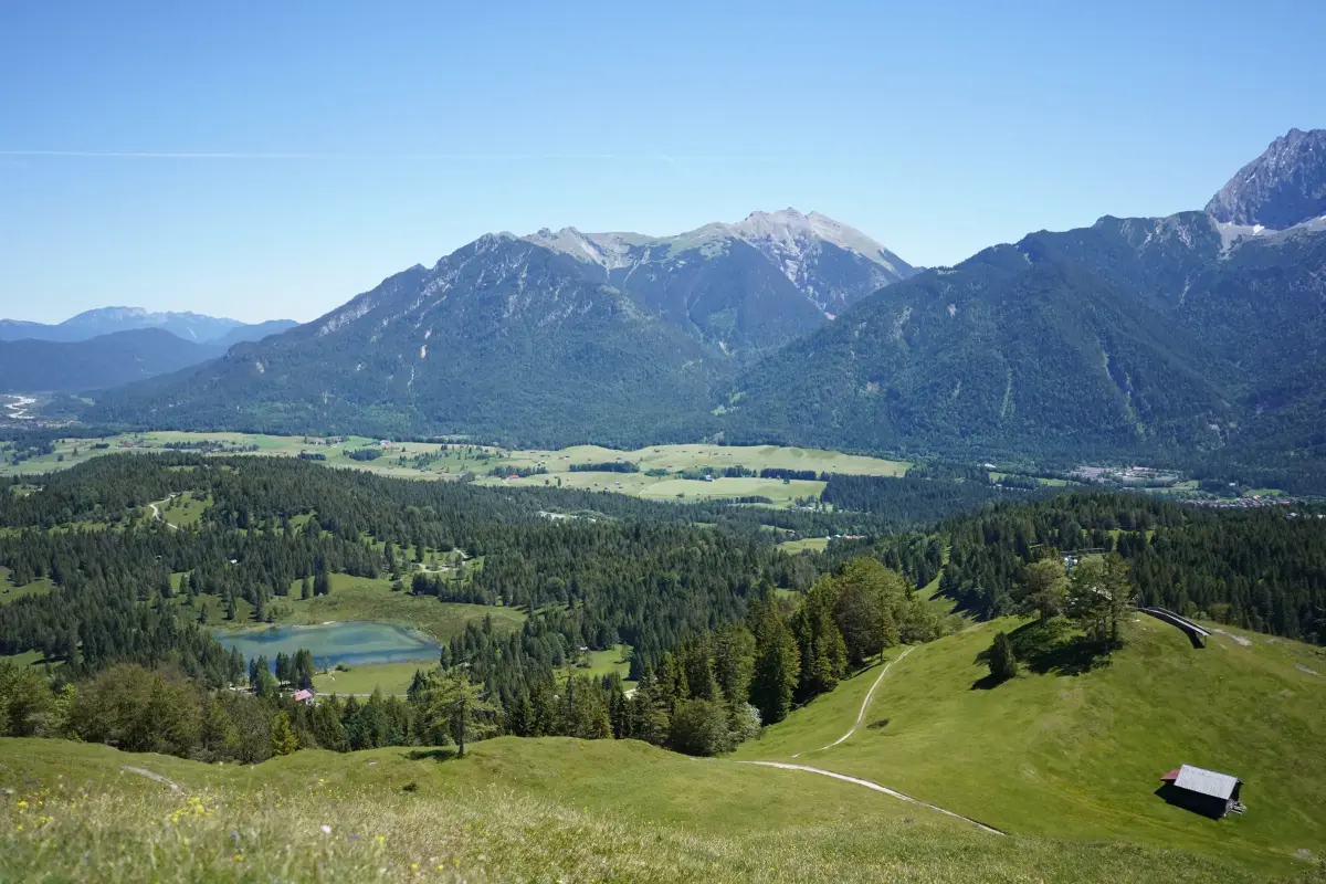 Blick vom Berg aus ins Tal mit Wäldern und einem See. Im Hintergrund Berge und blauer Himmel.