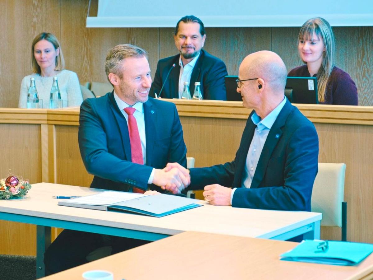 Zwei Männr im Vordergrund, die sich die Hand reichen. Im Hintergrund sitzen zwei Frauen und ein Mann und sehen zu.