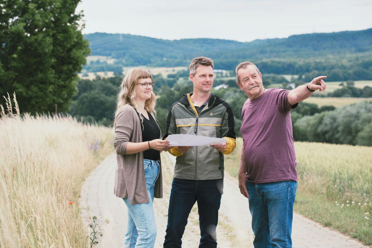 Drei Personen stehen auf einem Wirtschaftsweg. Eine Frau und ein Mann halten einen Plan in der Hand, ein weiterer Mann zeigt in die Landschaft.