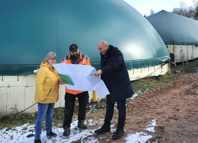 Zwei Männer und eine Frau stehen vor einer Biogasanlage und schauen auf einen Plan, den sie in der Hand halten.