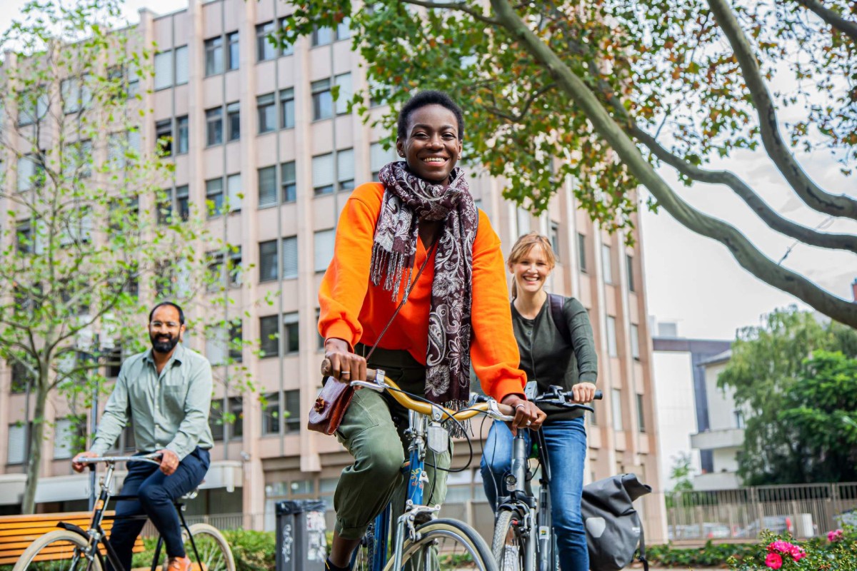 Drei Personen fahren in der Stadt Fahrrad. Im Hintergrund Bäume und ein mehrstöckiges Haus.