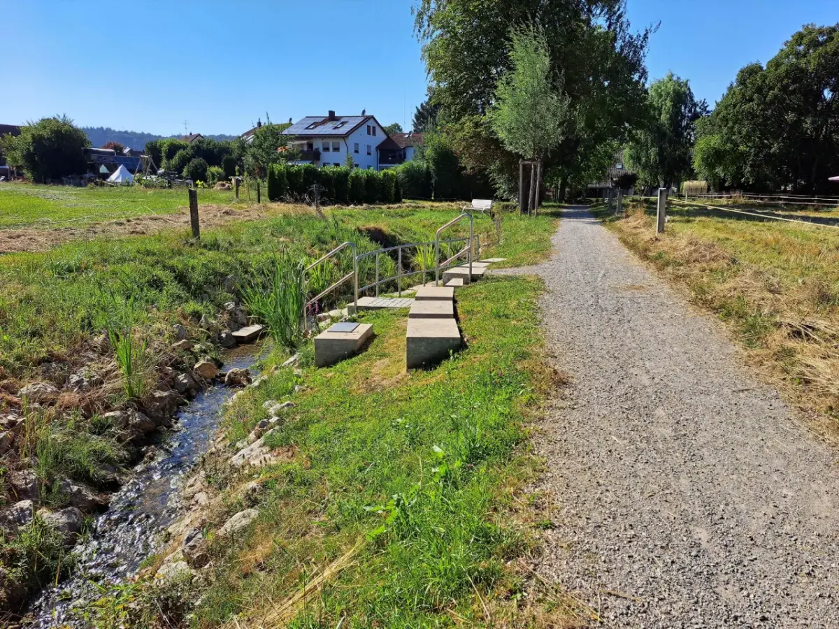Der renaturierte Mutterbach mit Kneipp-Anlage. Rechts ein Kiesweg. Im Hintergrund Häuser und Bäume bei gutem Wetter.