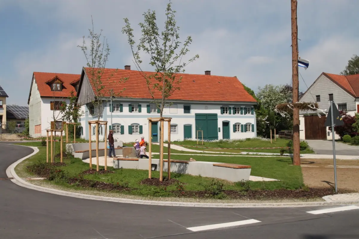 Der neugestaltete Dorfplatz mit Brunnen, Sitzbänken und neuen Bäumen. Häuser im Hintergrund. Leicht bewölkter Himmel.