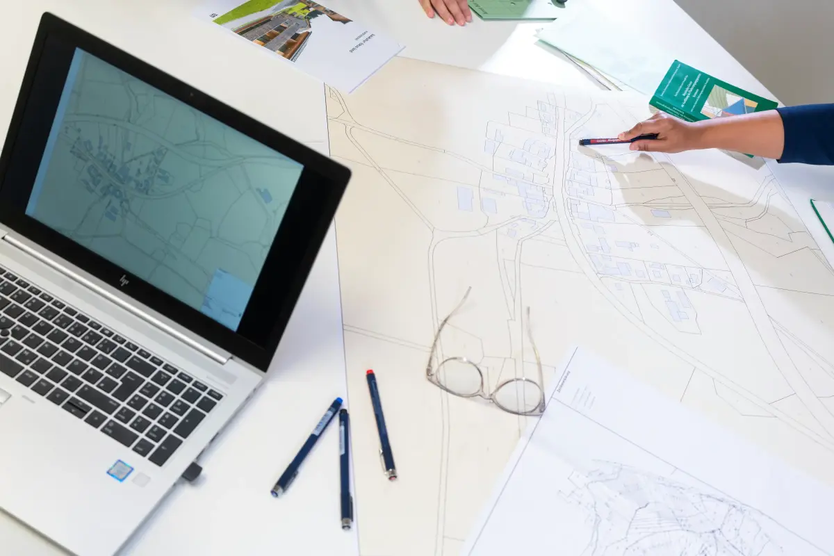 Ein Besprechungstisch ist mit großformatigen Karten bedeckt. Ein Laptop, Aktenmappen und Bücher liegen ebenfalls auf der das Bild füllenden, hellen Tischplatte. Rechts ragt eine Hand ins Bild und zeigt mit dem Stift auf ein Kartendetail.