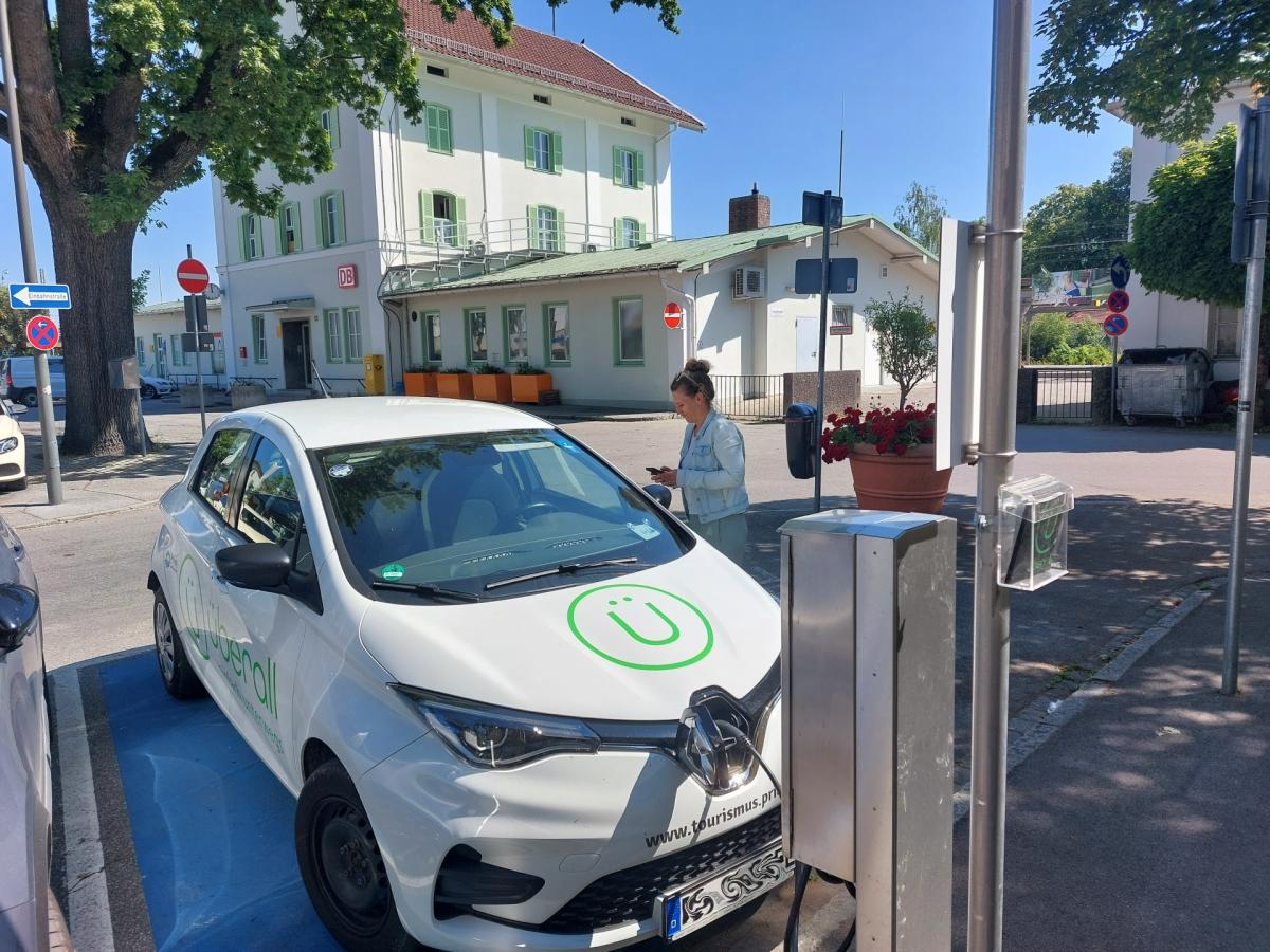 Frau steht rechts von weißem Carsharingauto. Im Hintergrund Bahnhofsgebäude.