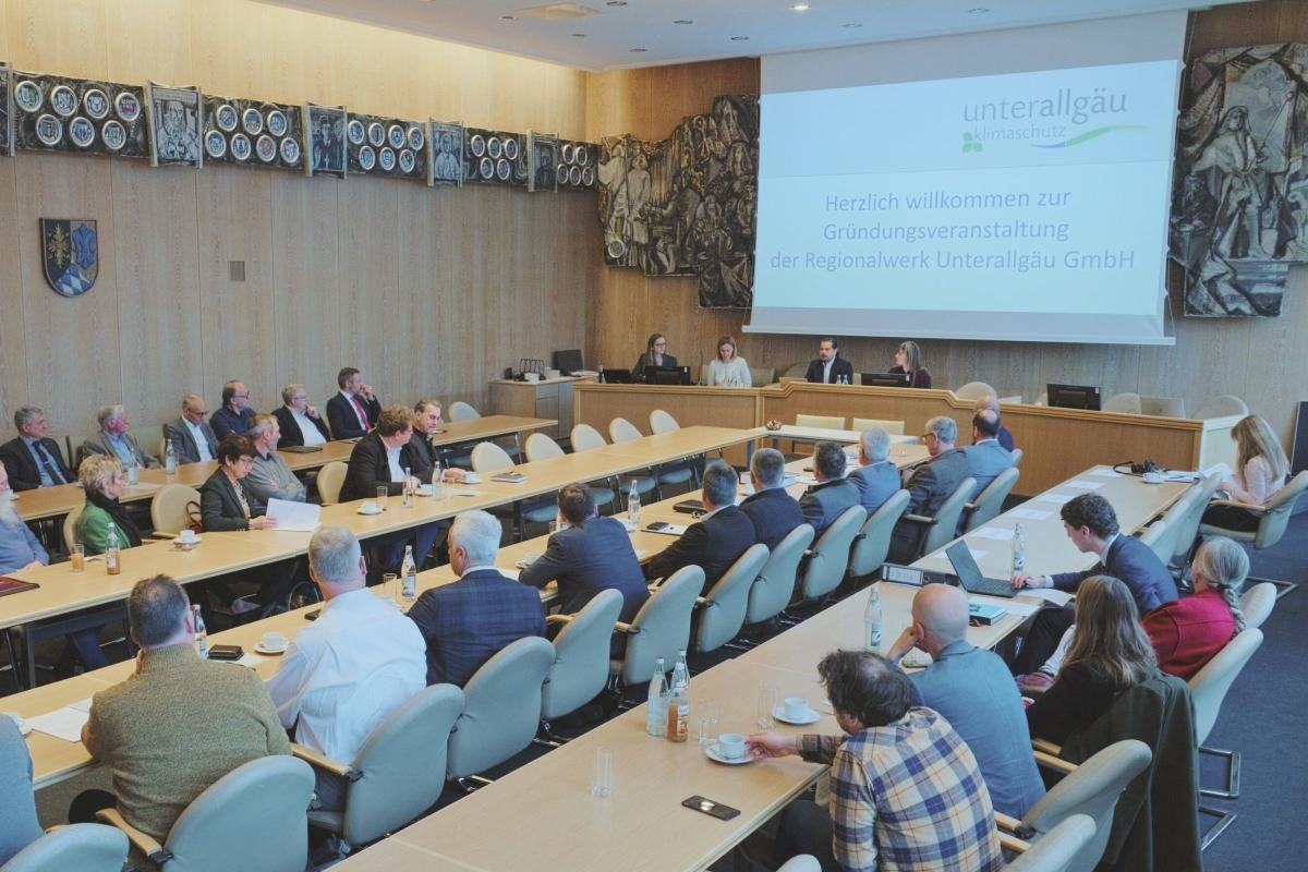Mehrer Menschen sitzen in einem Saal an vier langen Tischen und schauen zu der Power Point ant der Wand rechts oben.