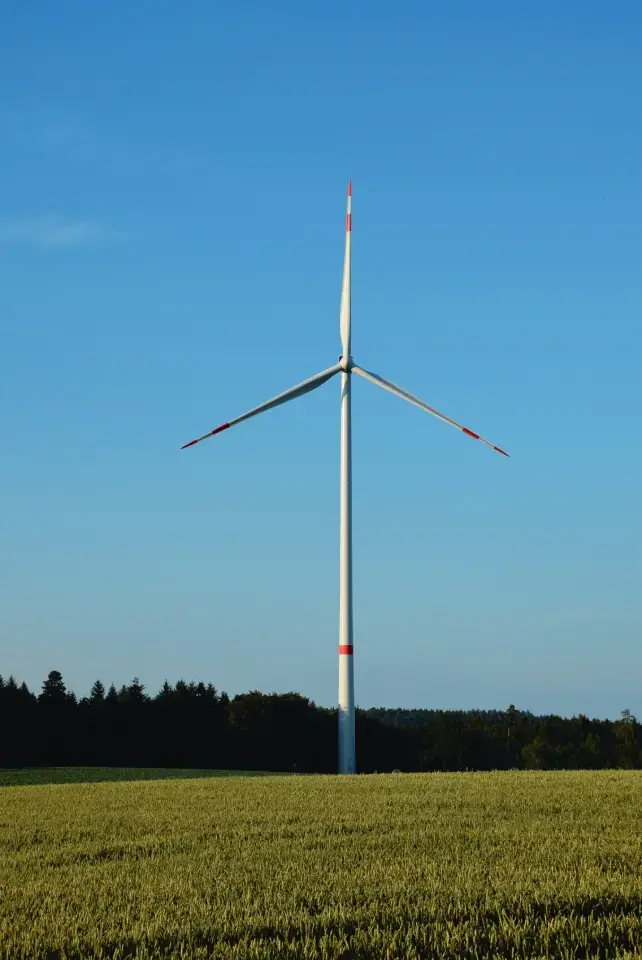 Windrad stehl am Waldrand. Davor Feld. Im Hintergrund ein Wald un blauer, strahlender Himmel.