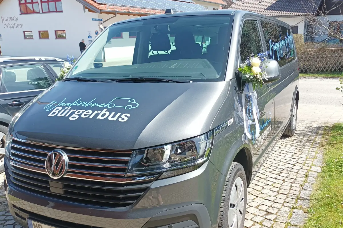 Grauer Bürgerbuss mit Blumengesteck am Spiegel. Links ein schwarzes Auto. Im Hintergrund ein Feuerwehrhaus und ein Wohngebäude.