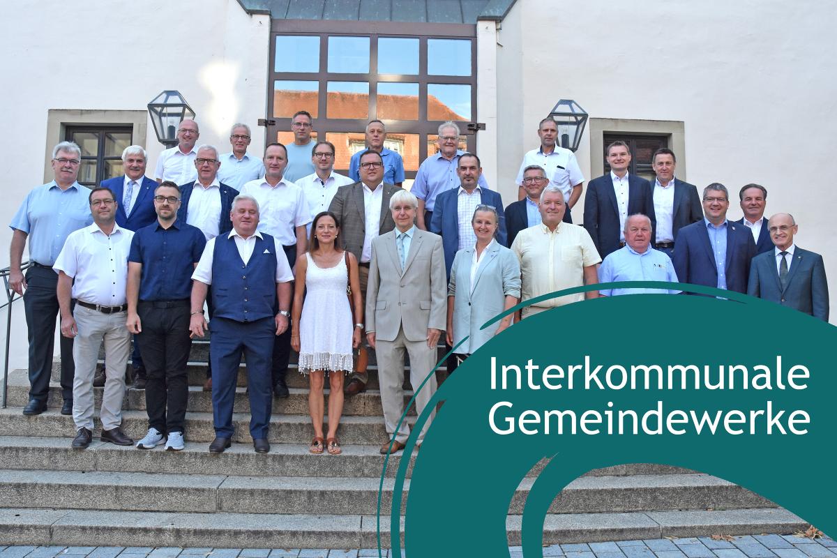 Personen stehen auf einer Treppe uns sehen lächelnd in die Kamera. Im rechten unteren Eck blauer Farbklecks mit Schriftzug "Interkommunale Gemeindewerke"