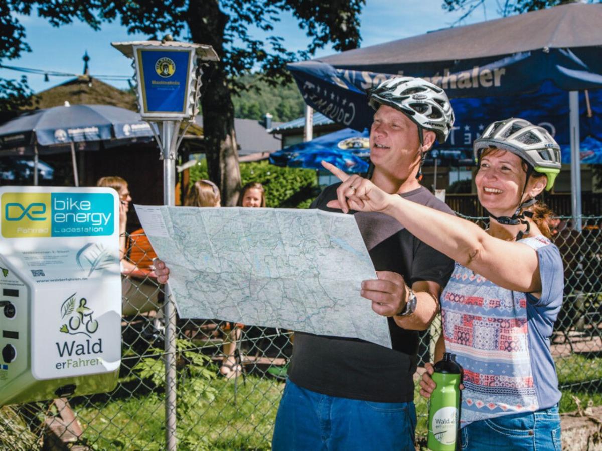 Man und Frau mit Fahrradhelm halten eine Karte und zeigen in die gleiche Richtung. Links die E-Bike-Ladestation.