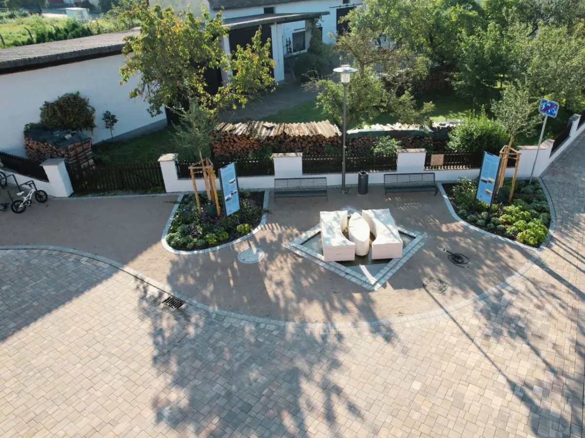 Sitzgelegenheit auf gepflastertem Platz. Links und rechts davon Baumpflanzungen und Begrünung. Im Zentrum ein Wasserspiel.