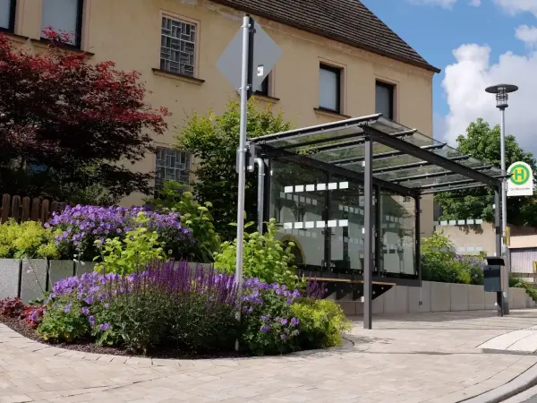 In der Mitte ein Bushäusschen. Im Hintergrund ein Haus. Im Vordergrund ein Blumenbeet und ein gepflasterter Gehweg.